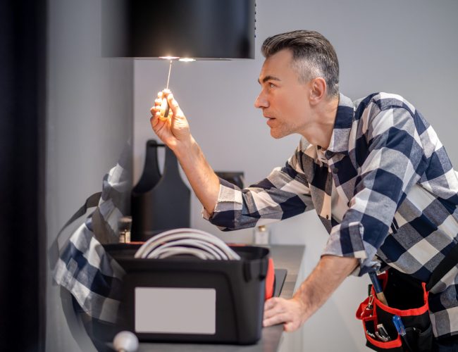 Profile of man touching lamp on kitchen hood Renovation. Profile of concentrated middle aged man in plaid shirt touching screwdriver to lamp on kitchen range hood in modern kitchen at home