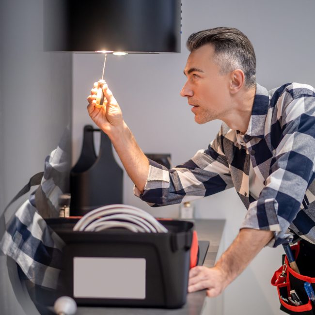 Renovation. Profile of concentrated middle aged man in plaid shirt touching screwdriver to lamp on kitchen range hood in modern kitchen at home
